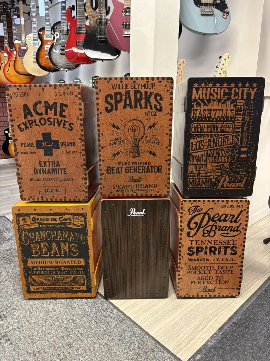 Six cajóns Pearl empilés, aux caisses sérigraphiées façon vieilles caisses de bois, devant un mur de guitares électriques.