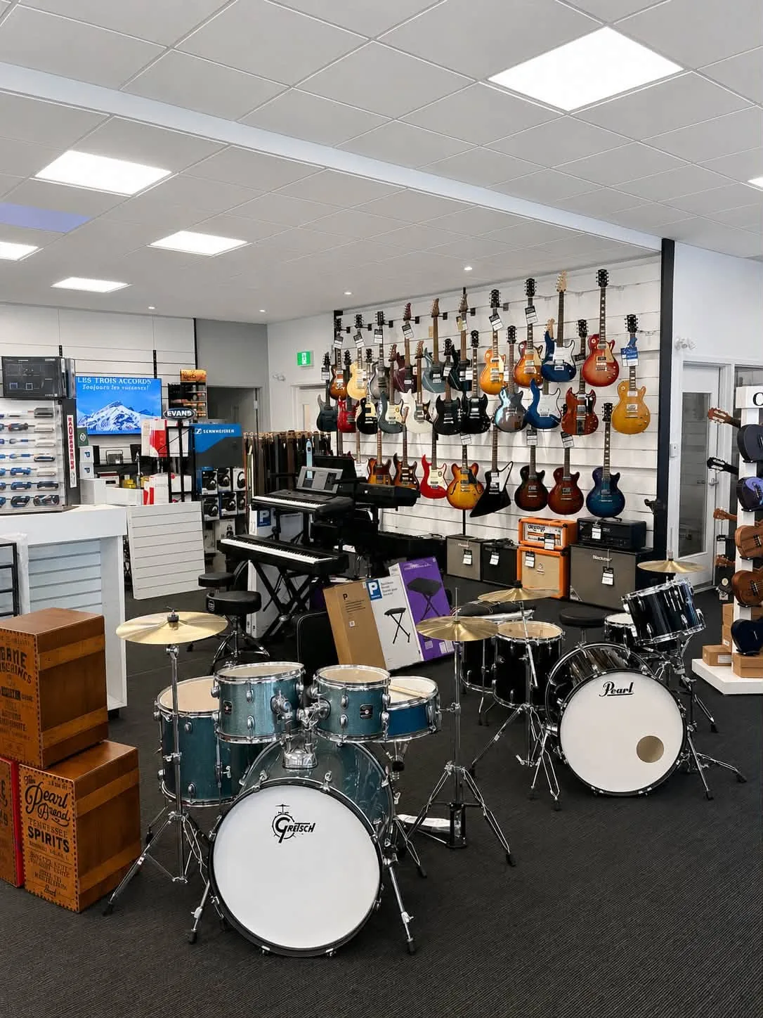 Vue d’ensemble du magasin Multi Musique : mur de guitares électriques, claviers, deux batteries montées, amplis et présentoirs d’accessoires.