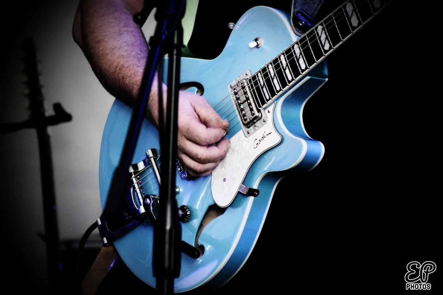 Gros plan d’une guitare électrique Godin bleu pâle jouée sur scène, sous un éclairage sombre, devant un pied de micro.